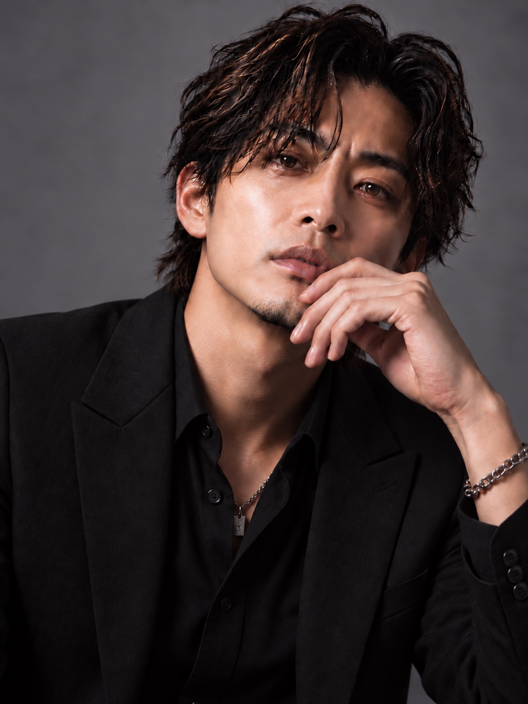 グレーの背景で黒いシャツとジャケットを着て、顔に手を添えるDAISUKE